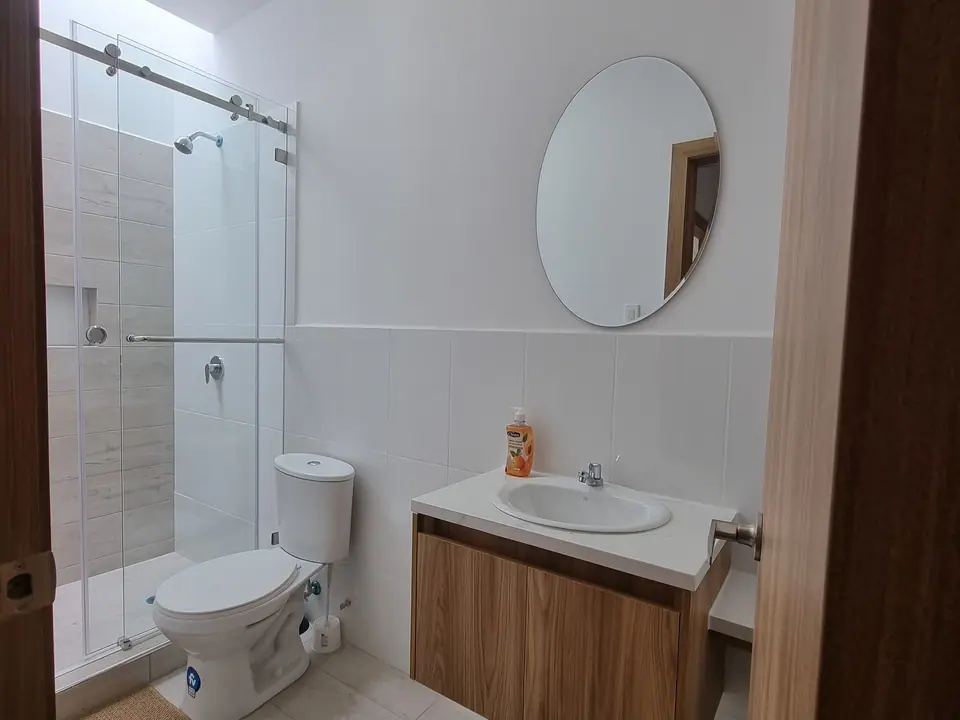Un baño con ducha de puerta de vidrio, lavamanos sobre mueble de madera y espejo redondo.