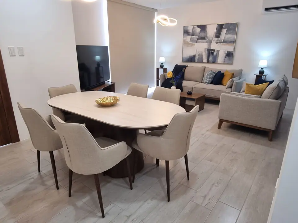 El comedor visto hacia la sala: la mesa ovalada, seis sillas tapizadas y el televisor.