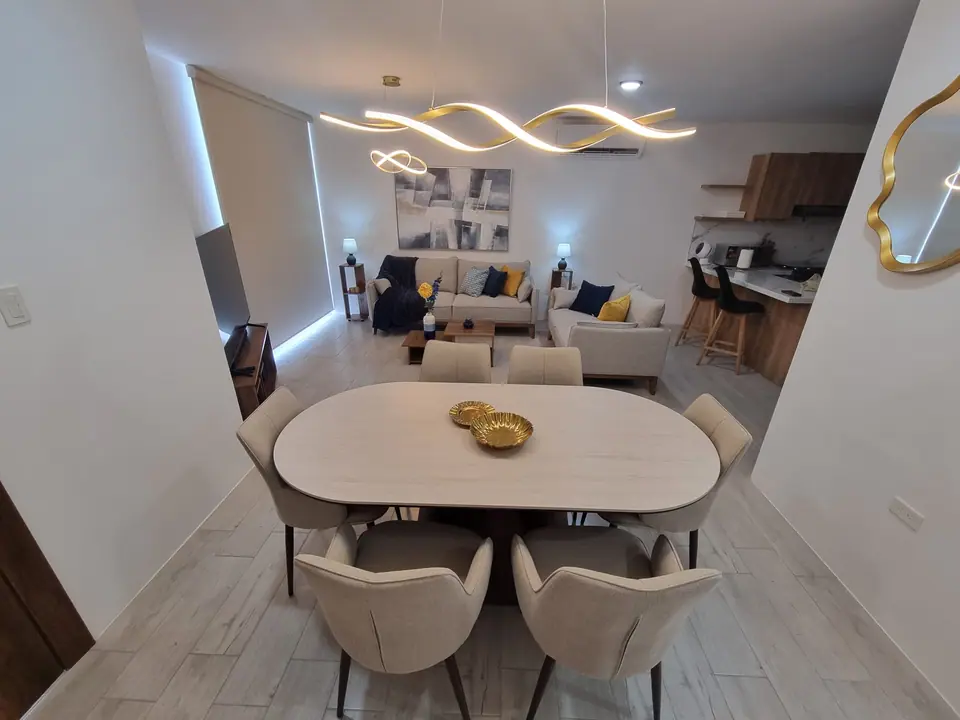 La mesa de comedor para seis bajo una lámpara dorada, con la sala y la cocina al fondo.