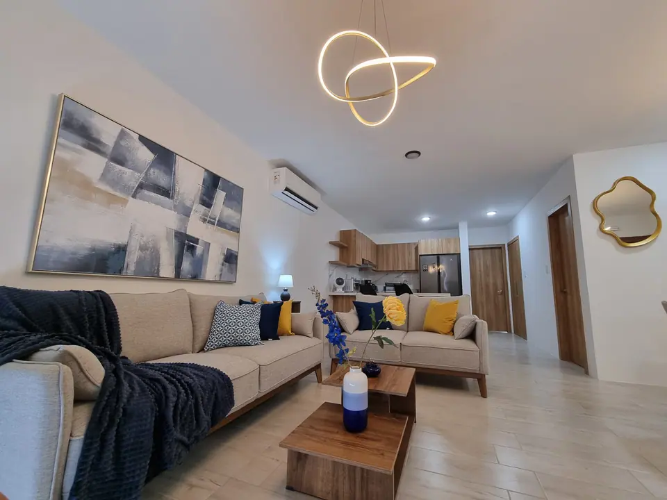 La sala vista desde la entrada: un sofá largo con cojines azules y amarillos, un cuadro abstracto y una lámpara dorada.