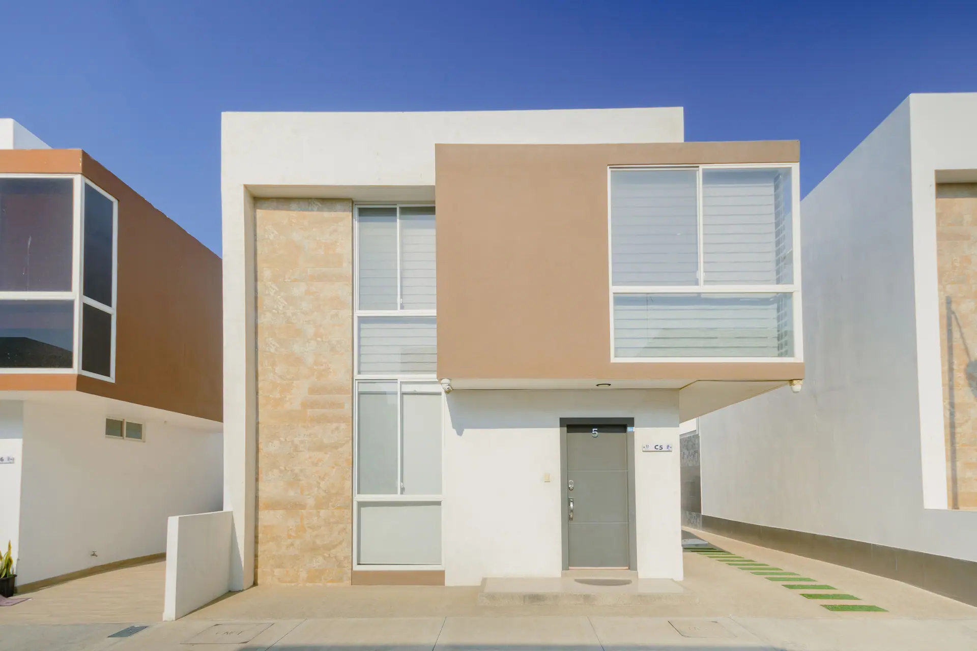 Fachada de CASA C5: casa blanca de dos plantas con un panel de piedra clara y un volumen color arena.