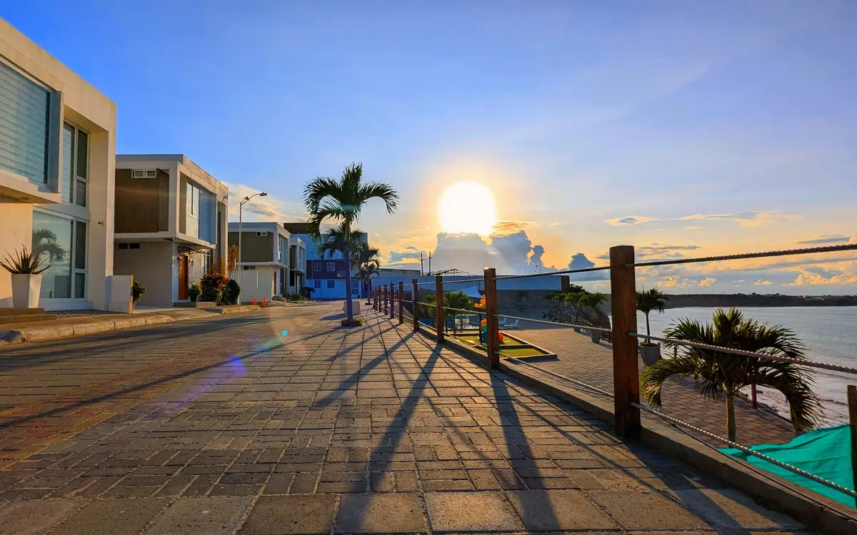 El malecón de la urbanización al atardecer: palmeras, barandal de madera y el sol sobre la bahía.