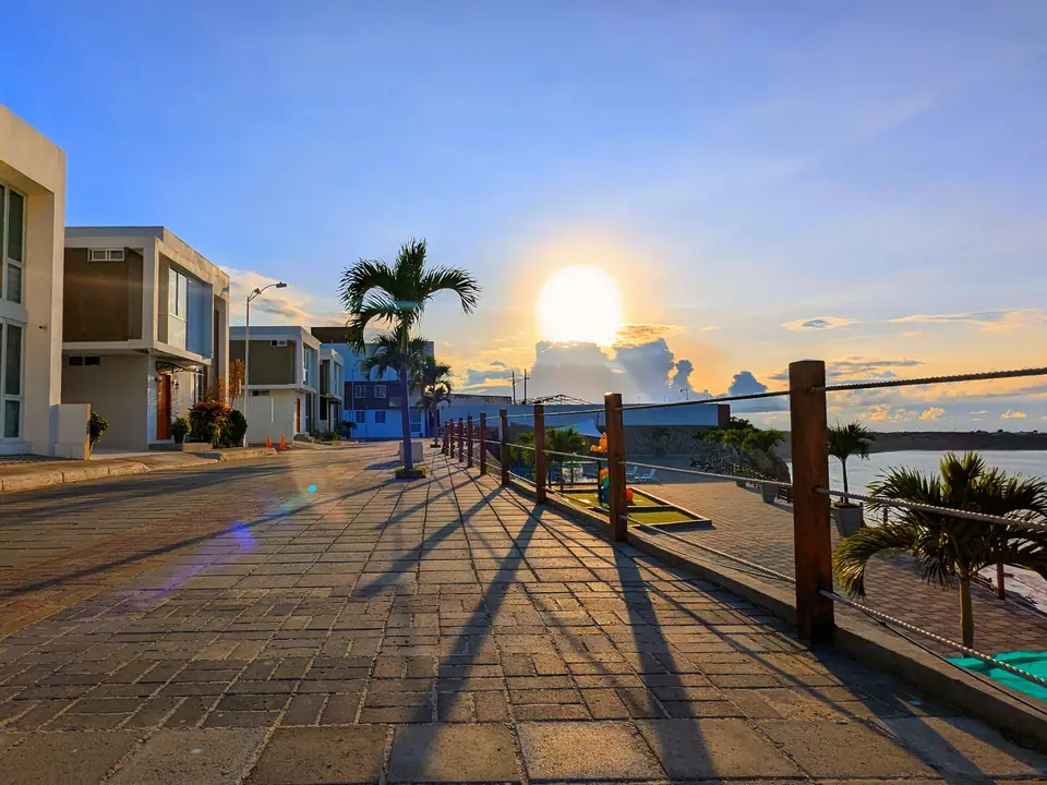 El malecón de la urbanización al atardecer: palmeras, barandal de madera y el sol sobre la bahía.